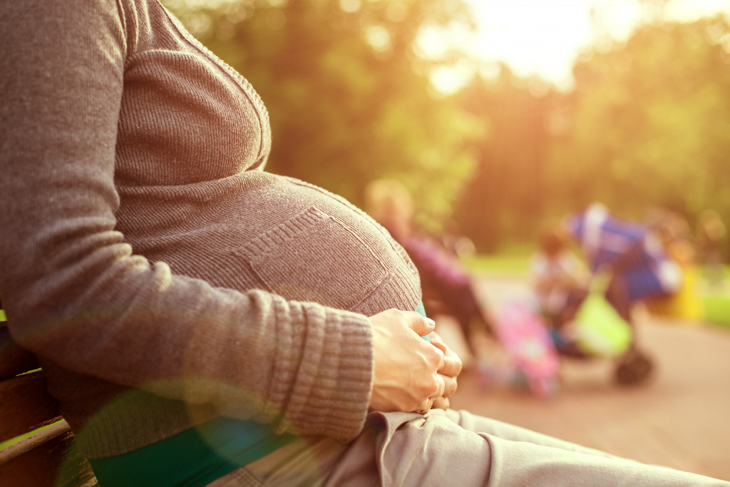 In a park, a pregnant woman is sitting on a bench in a long-sleeved top.